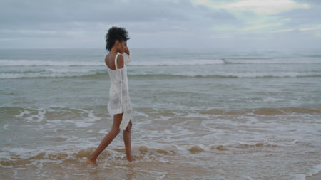 Sad girl walking ocean beach in white bikini on gloomy day. Thinking curly lady going splashing waves contemplating life personal problems at sea shore. Upset african american woman going rest alone.の写真素材