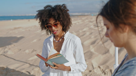 Focused girl reading book at ocean picnic. Two friends resting on sandy beach in summer sunlight. Attractive african american studying enjoying sea breeze. Love partners spending time relax on coast.の写真素材