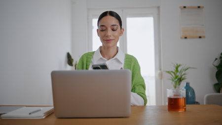 Smiling businesswoman picking up smartphone at computer table remote office closeup. Laughing student girl talking mobile phone reacting on jokes. Young lady gossiping cellphone having fun at houseの写真素材