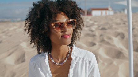 Stylish woman enjoying beach on sunny day closeup. Happy african american girl nod communicate on sandy seashore. Attractive curly lady relax outdoors wear sunglasses. Summer weekend leisure conceptの写真素材