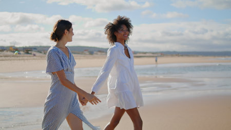 Joyful lesbian couple walking sea shore on sunny day. Happy girls holding hands resting on ocean coastline. Multicultural best friends talking spending weekend together. Woman friendship love conceptの写真素材