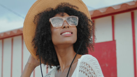 African american enjoying sunlight at beach house closeup. Stylish curly girl wearing sunglasses hat resting at ocean shore. Smiling tourist spending summer vacation at coast. Romantic mood conceptの写真素材
