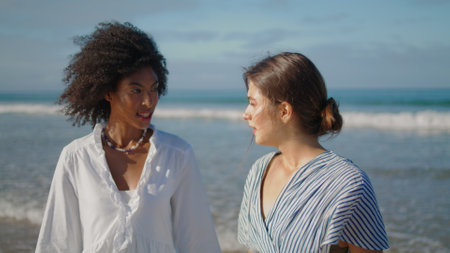 Stylish girls talking beach in summer sunlight closeup. Smiling lgbt couple rest sharing secrets at ocean waves. Beautiful multicultural women spending weekend at shore. Bonding friendship concept.の写真素材