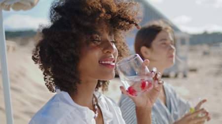 Beautiful woman enjoying cocktail closeup. Happy african american drinking soda on summer picnic. Satisfied curly girl resting with girlfriend love partner at sea shore. Carefree idyllic morning.の写真素材
