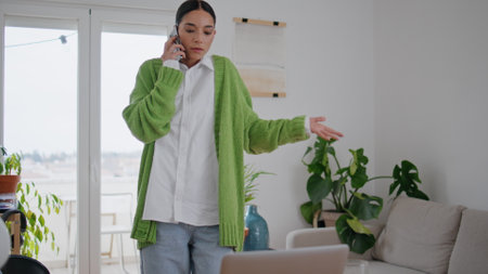 Gesturing businesswoman calling mobile phone walking at room. Confused woman communicating cellphone alone at cozy white apartment interior. Focused lady talking telephone solving work moments indoorの写真素材