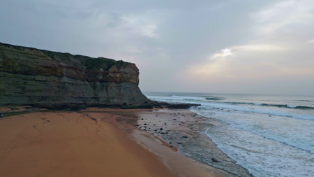 Gloomy marine beach landscape at cloudy day. Panoramic serene sandy coast with calm ocean waves slow motion. Grey sea water surface washing seaside with volcanic cliffs background. Stormy wild sceneryの写真素材