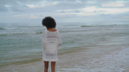 Woman contemplating ocean beach in white outfit. Curly slim girl looking sea thinking of personal problems loneliness. Unrecognized romantic african american walking waves coast on gloomy day alone.の写真素材