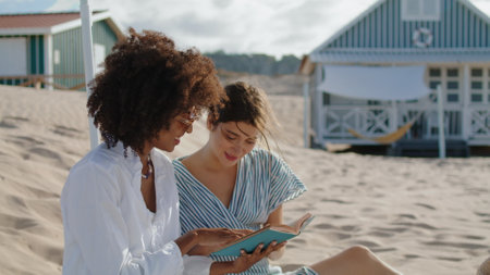 Two girls reading book on summer beach picnic. Happy lesbian couple talking enjoying novel in sunlight. Carefree multiethnic friends discussing literature on sandy coast. Vacation weekend concept.の写真素材