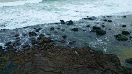 Foaming surf covering rocky coastline closeup. White ocean waves beating of volcanic pebble beach slow motion. Sea breaking along shore. Seabirds flying water splashing sand coast. Nature backgroundの写真素材