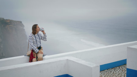 Relaxed tourist drinking coffee at beautiful seaside view. Girl enjoy vacation resting alone on mediterranean white rooftop. Beautiful street photographer admire scenic view. Peaceful trip morning.の写真素材