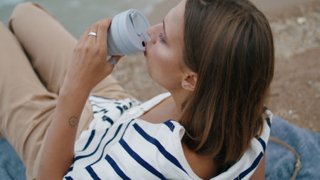 Woman drinking picnic coffee on cliff edge closeup. Pensive girl rest seashore enjoying view on mountain top. Serene tourist relax taste morning beverage in eco friendly cup. Holiday chillout conceptの写真素材