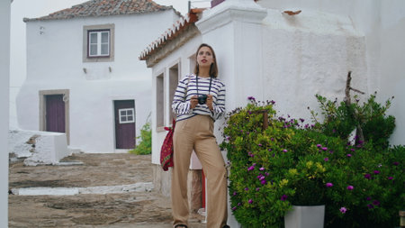 Tourist admiring city architecture on vacation. Pretty girl posing narrow street leaning white houses in summer. Beautiful dreaming woman hold analog camera resting in old town. Hobby travel conceptの写真素材