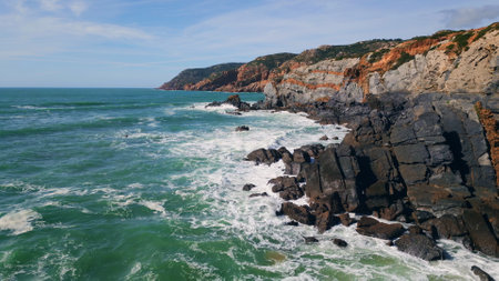Ocean surf splashing on coastal rugged cliffs with white foam super slow motion. Aerial view stormy beautiful sea waves breaking on volcanic dark rocky seashore. Amazing marine landscape sunny day.の写真素材