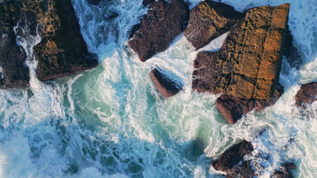 Beautiful stormy sea foam splashing at seashore cliffs in super slow motion. Picturesque foamy ocean waves crashing on dark coastal rocks sunny day. Aerial view rough seashore stones washing by water.の写真素材