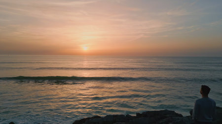 Man silhouette watching sunset at dark cliffs back view. Panoramic dawn marine landscape drone view. Endless scenic ocean horizon. Pastel pink skyline. Orange sunset reflecting in calm lapping waterの写真素材