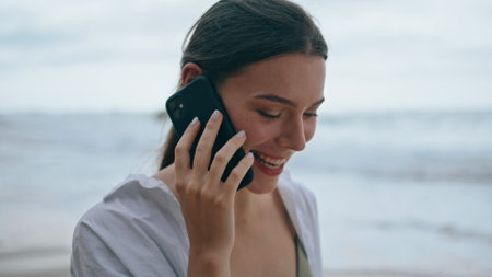 Smiling cheerful girl calling phone on gloomy beach closeup. Portrait of attractive happy woman talking by modern smartphone walking empty seashore. Beautiful lady enjoy mobile communication on natureの写真素材