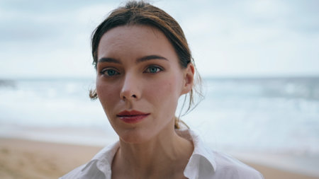 Beautiful gentle woman face looking camera intently in front sea waves closeup. Portrait of attractive pensive girl standing alone at cloudy beach wearing white shirt. Tranquil lady posing on seashoreの写真素材