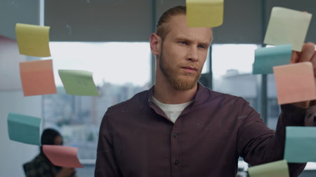 Thoughtful man pondering sticker wall office closeup. Young freelancer preparing tasks on sticky notes transparent board. Serious startuper brainstorming creative ideas at big windows coworkingの写真素材