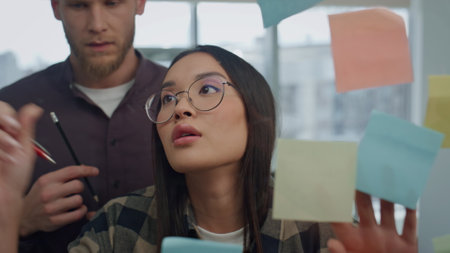 Eyeglasses creator writing sticker wall at office close up. Asian woman talking to man colleague sharing ideas together. Involved startupers team using sticky notes for brainstorming session at homeの写真素材