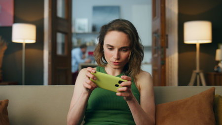 Focused gamer playing mobile phone at home closeup. Carefree woman enjoying smartphone video games relaxing at sofa weekend. Involved lady sitting couch entertaining at house zoom out. Girl having funの写真素材