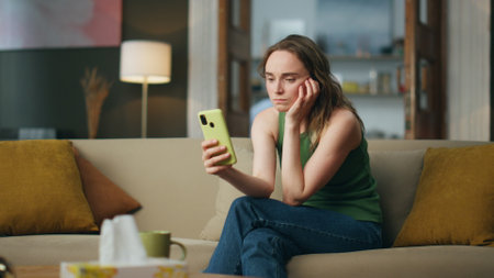 Sad lady using smartphone on home sofa. Depressed woman talking mobile phone video chat at living room. Unhappy girl videocalling cellphone sitting couch. Annoyed person listening interlocutor indoorsの写真素材