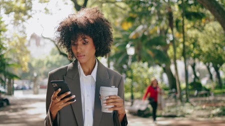 Confident businesswoman calling standing city park with paper coffee cup close up. Curly attractive african american girl using phone wearing elegant suit. Stylish woman holding mug takeaway drink.の写真素材