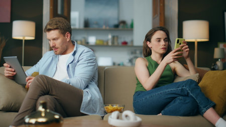 Involved couple reading devices at living room. Bearded focused man watching tablet. Relaxed woman browsing mobile phone at cozy sofa. Internet addicted family using gadgets at comfort house togetherの写真素材