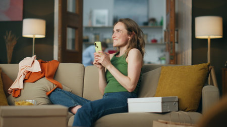 Emotional girl rejoicing mobile phone at home. Shopping addicted person sitting sofa ordering new items. Happy smiling woman dancing hands with smartphone relaxing around boxes and clothes aloneの写真素材