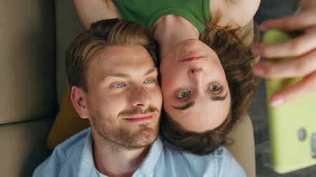 Cute lovers photographing smartphone chilling sofa closeup. Happy woman making selfie laying couch with smiling husband top view. Loving pair resting together at home. Lazy domestic weekend conceptの写真素材