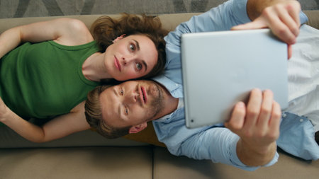 Funny couple taking selfie laying couch top view. Happy man photographing tablet computer chilling sofa closeup. Cute woman showing tongue. Loving pair resting together at home apartment. Lazy weekendの写真素材