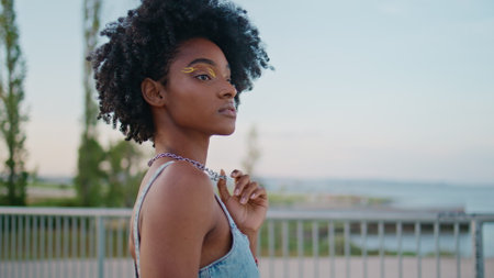 African teen standing street looking around close up. Beautiful curly model posing city street playing with jewelry on neck. Portrait of gorgeous young lady with bright makeup relaxing in evening townの写真素材