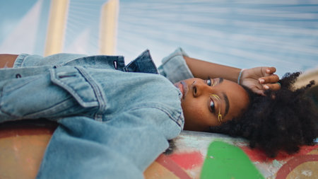 Stylish model posing lying outdoors in jeans jacket close up. Beautiful african american girl with bright makeup relaxing at urban street alone. Trendy black haired hipster looking camera confidently.の写真素材