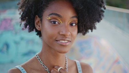 Charming curly girl smiling posing at skate park close up. Gorgeous african american teenager looking camera enjoying city leisure. Happy carefree female teen with modern makeup relaxing outdoors.の写真素材