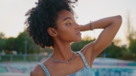 African american hipster posing at urban street close up. Beautiful glamorous girl with bright makeup standing summer city park alone. Stylish young woman looking camera touching curly hairstyle.の写真素材