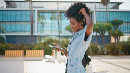 Joyful teen girl messaging on smartphone walking sunny city close up. Happy african american teenager smiling reading sms on cellphone. Cheerful curly hipster laughing watching social media on phone.の写真素材