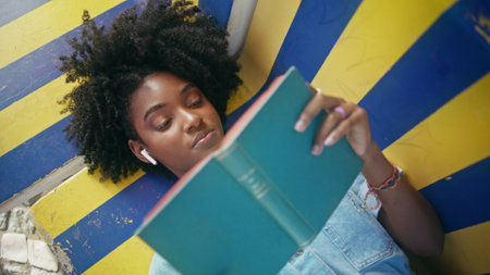 Closeup teen reading book aloud lying on colorful bench outdoors. Curly african american girl in headphones enjoy literature hobby. Beautiful young woman relaxing with favorite novel at summer weekendの写真素材