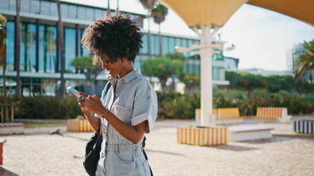 Smiling woman texting at mobile phone on go sunny day close up. Happy african american girl messaging looking smartphone screen on summer walk. Cute curly lady enjoy remote communication outdoors.の写真素材