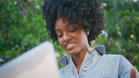 Black-haired woman videocalling by laptop sitting under green trees closeup. Portrait of beautiful smiling african american girl talking to computer web camera outdoor. Cute freelancer speaking onlineの写真素材