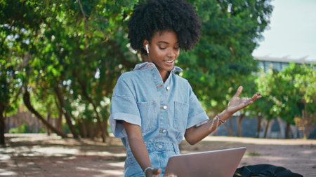 Teenager speaking at video call by laptop sitting in green park close up. Attractive african american girl in wireless earbuds talking to computer web camera on street. Curly woman chatting online.の写真素材