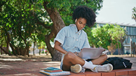 Curly teenager studying online with laptop sitting sunny street. Busy african american girl searching internet information making notes. Smart young woman writing synopsis from computer screen outdoorの写真素材
