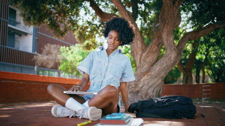 Curly woman closing laptop stretching neck after remote work on sunny nature. Tired african american girl relaxing after online studying in green park. Black haired teen sitting street with notebook.の写真素材