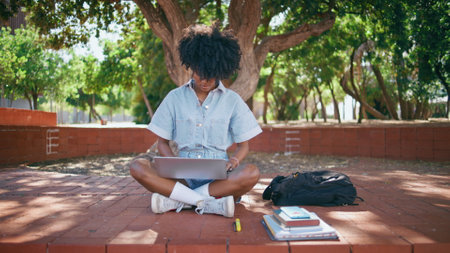 Curly student looking laptop screen sitting city park alone. Focused african american young woman studying online using notebook outdoors. Black-haired teen girl searching information in internet.の写真素材