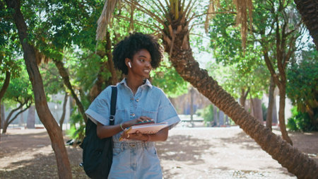 Teen girl walking campus college holding books sunny day close up. Cute african american student greeting with friends in green park. Beautiful female teenager with backpack going on university lessonの写真素材