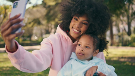 Mother making phone selfie with cute little daughter at green park close up. Happy african american woman taking photo sitting on meadow with curly child. Cheerful girls smiling to smartphone camera.の写真素材