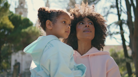Mom holding adorable daughter walking on sunny street close up. Carefree african american woman communicating with cute little girl on nature stroll. Happy curly mother carrying pretty child on hands.の写真素材