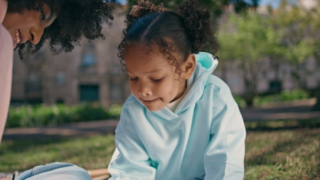 Happy child drawing on picnic sunny day with smiling mother close up. Cute little girl enjoy creativity holding pencil outdoors. African american young woman watching on daughter hobby sitting meadow.の写真素材