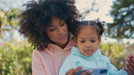 African american child watching cartoon at telephone in mom hands close up. Joyful young mother showing entertaining mobile app to little daughter on nature. Carefree happy family enjoy sunny weekend.の写真素材
