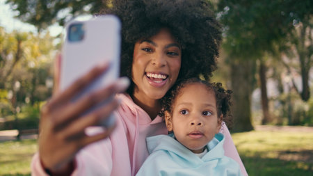 Mother child taking photo together on phone at beautiful green meadow close up. Smiling cheerful african american woman making selfie with little daughter using modern smartphone. Technology concept.の写真素材