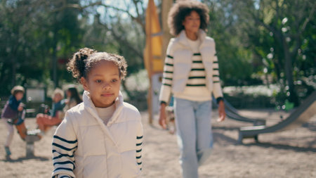 Child walking playground with mom enjoying carefree sunny weekend close up. Happy african american girl strolling park wearing stylish white jacket. Caring mother looking on lovely daughter outdoors.の写真素材