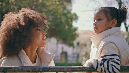 Mother looking on daughter with tenderness relaxing outdoors close up. Happy african american family spending weekend together in sunny park. Curly mom talking with adorable little girl on nature.の写真素材